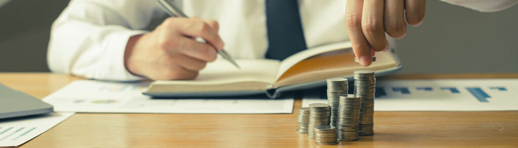 imagem de um homem engravatado anotando em um livro ao mesmo tempo que segura moedas para ilustrar texto que fala de correção monetária